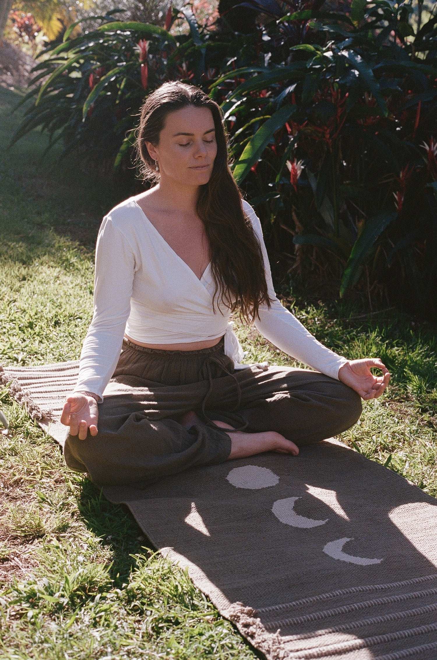 Woman meditating on a hand woven herbal dyed yoga mat outdoors with greenery in the background